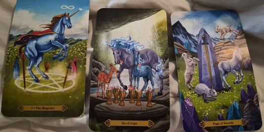 Three tarot cards laid out for the daily collective reading: The Magician, Six of Cups, and Page of Swords from a unicorn tarot deck
