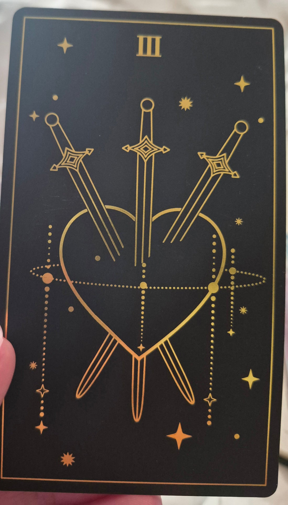 Three of Swords tarot card — gold swords piercing a heart on a black background with stars
