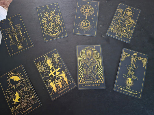 Eight tarot cards spread on a dark table — Seven of Cups, Ten of Pentacles, Two of Pentacles, Death, The Moon, Judgement, King of Swords, and The Hanged Man
